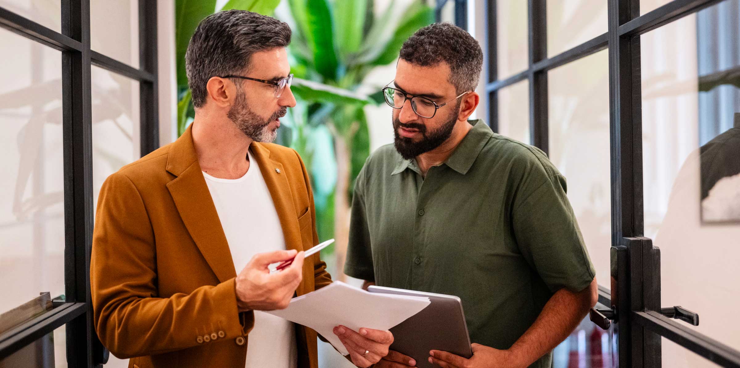 Two colleagues standing in a modern office hallway discussing documents, with one holding papers and a pen while the other holds a tablet; large green plants visible in the background.