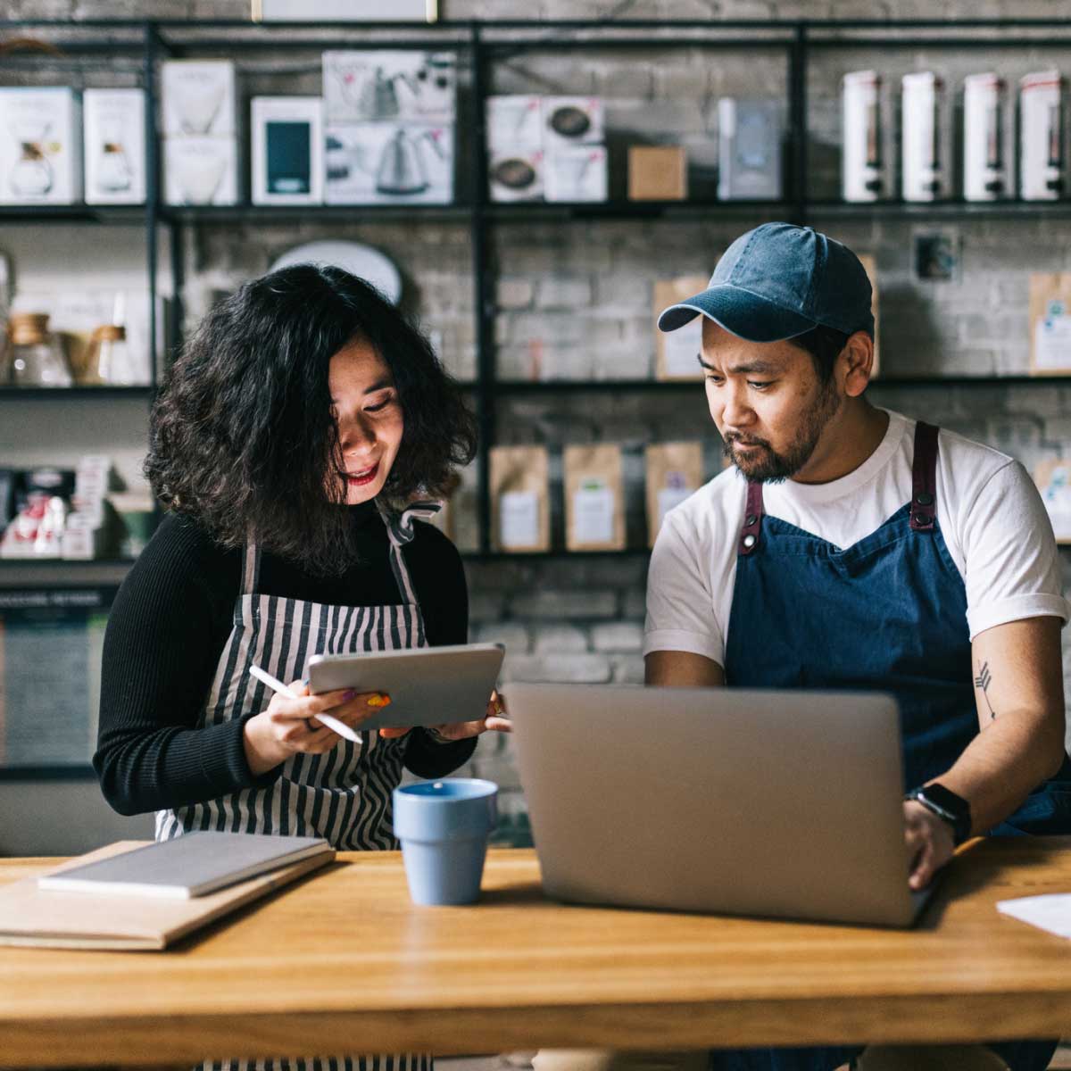 Two people in aprons working together at a café counter using a tablet and a laptop.