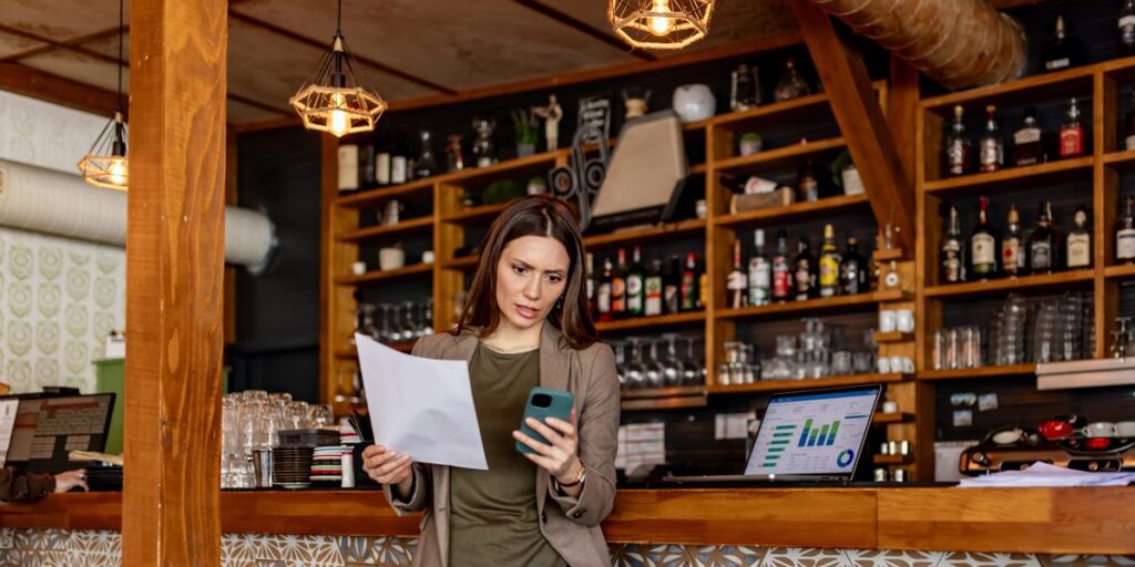 Woman at a café bar counter frowning at a printed invoice while checking her phone, a laptop dashboard beside her