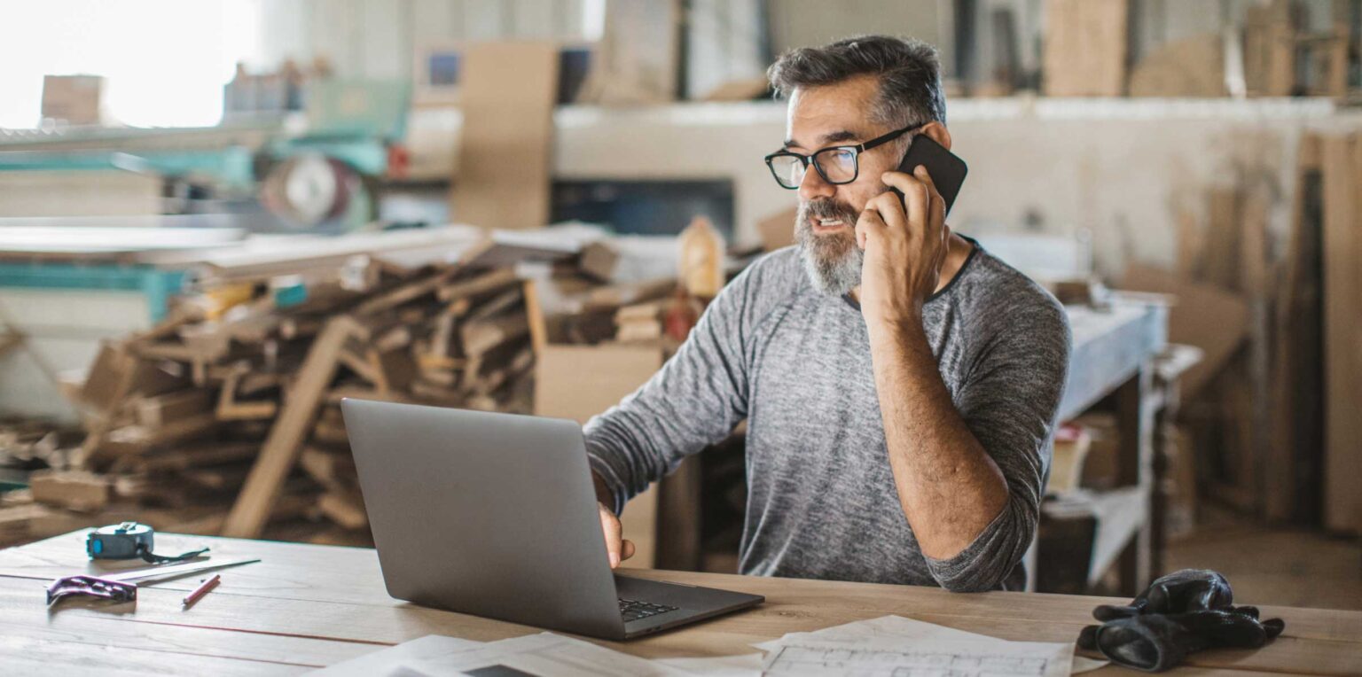 Carpenter on phone using laptop in workshop