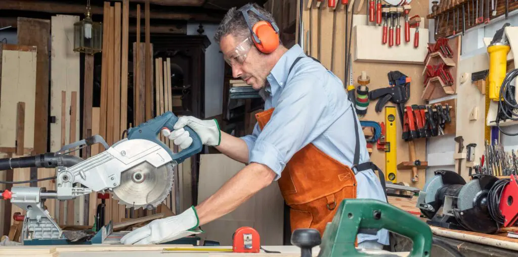 A woodworker in a workshop uses a compound mitre saw to cut timber on a workbench, wearing protective gloves, ear protection, and a leather apron, with tools and stacked wood visible in the background.