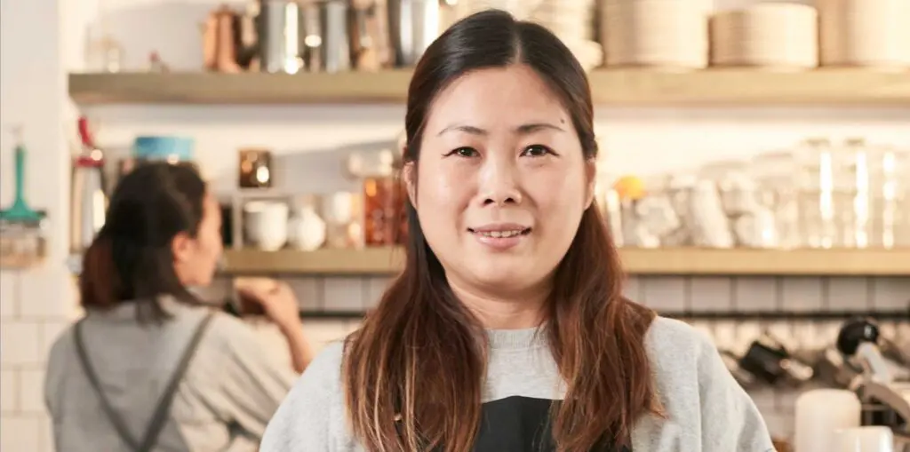 Cafe staff member wearing an apron holds a tablet behind the counter, with a busy coffee shop workspace in the background.