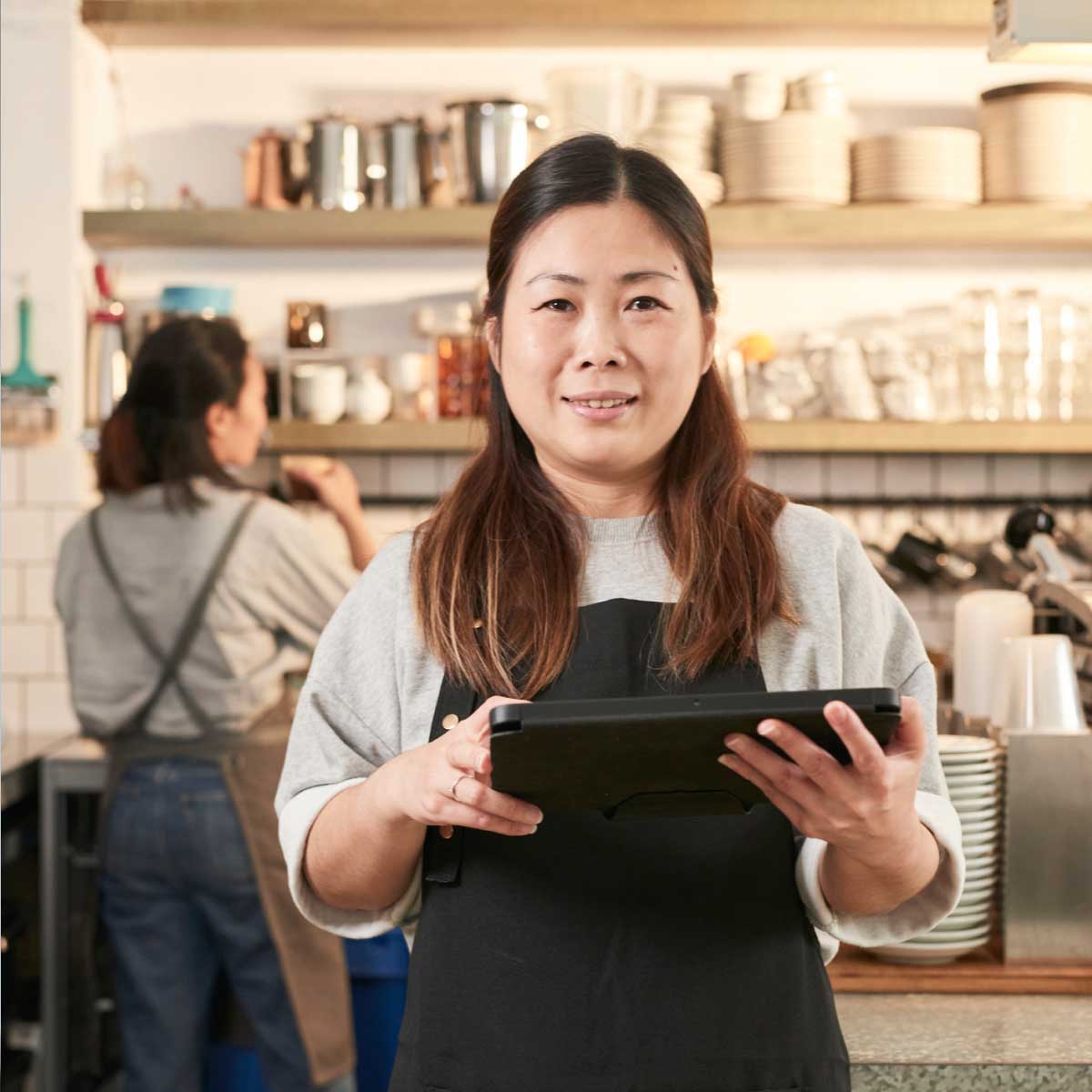 Cafe staff member wearing an apron holds a tablet behind the counter, with a busy coffee shop workspace in the background.