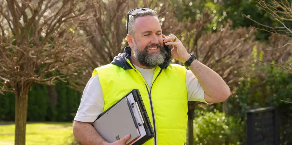 A person in a bright yellow safety vest stands outside among trees, holding a laptop and a measuring device while speaking on a mobile phone.