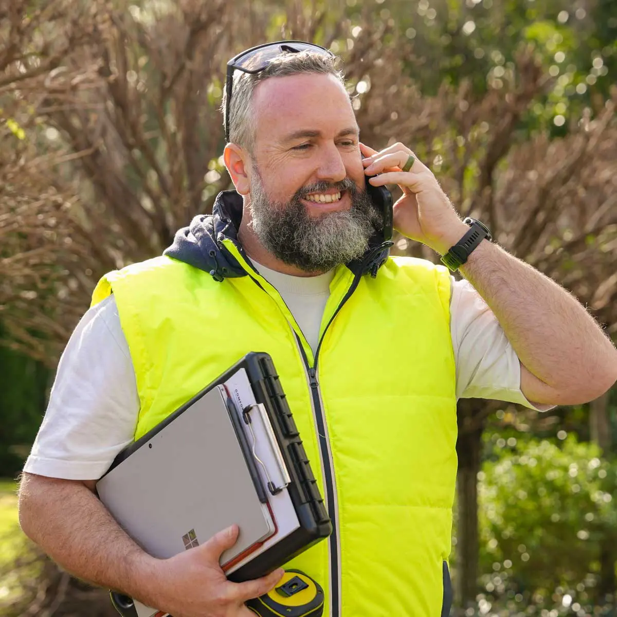 A person in a bright yellow safety vest stands outside among trees, holding a laptop and a measuring device while speaking on a mobile phone.