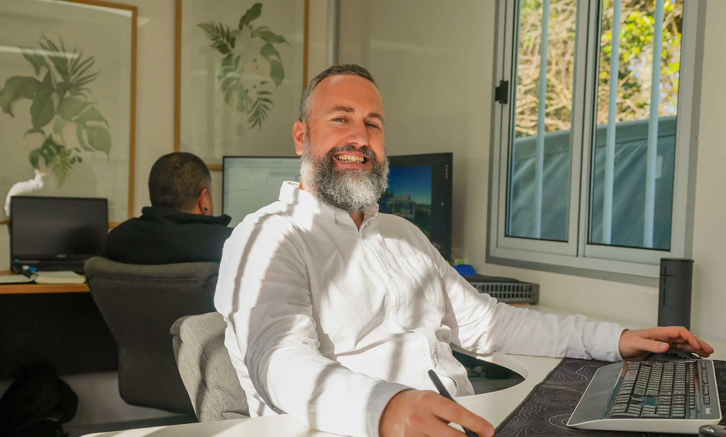 A person working at a desk in a bright office, using a computer while another person works in the background.