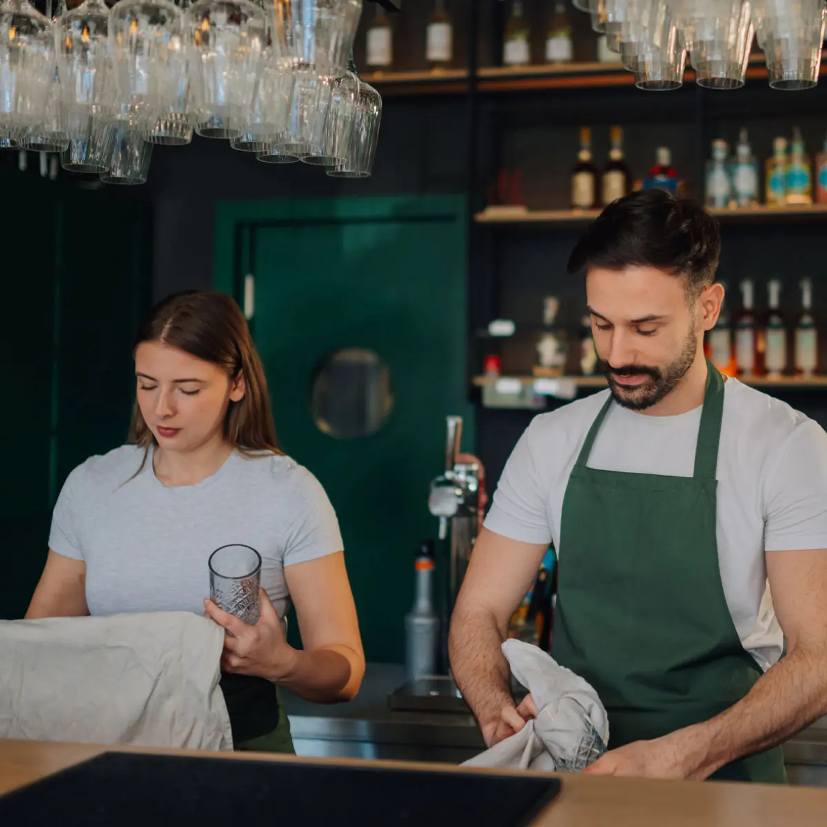 Two people working behind a bar with shelves of bottles and hanging glassware in the background.